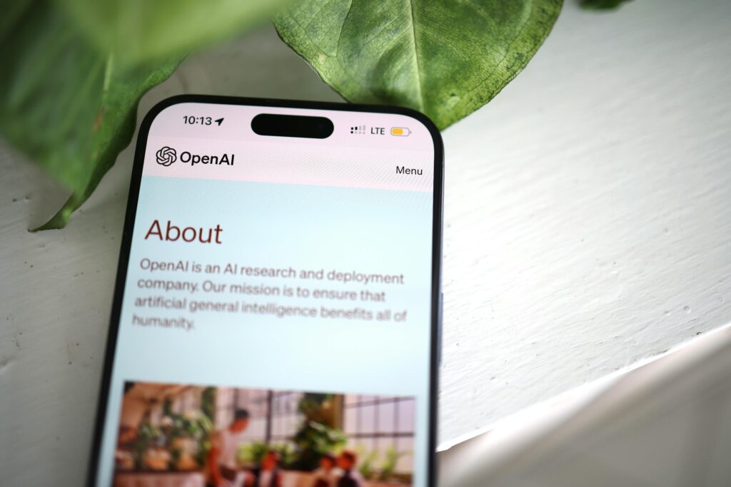 Current image: Close-up shot of a smartphone screen showing the OpenAI website with greenery in the background.